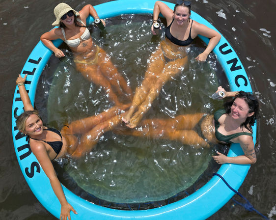 Aerial view of four friends in swimsuits lounging in a circular blue inflatable lake float with a clear center, legs meeting in the middle and each holding a canned drink on a sunny summer day.