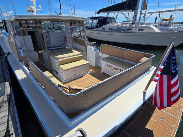 Sunlit motor yacht docked at a marina, open stern cockpit with tan cushioned seating, life jacket storage box, teak-style deck and an American flag with sailboats moored in the background.