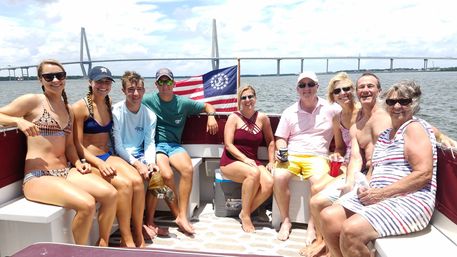 Multi-generation group enjoying a sunny pontoon-boat outing with an American flag, coastal water and a cable-stayed bridge in the background.