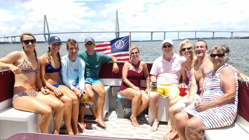 Multi-generation group enjoying a sunny pontoon-boat outing with an American flag, coastal water and a cable-stayed bridge in the background.