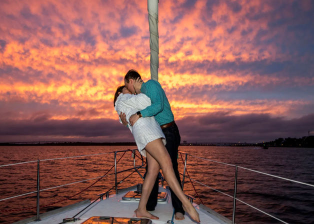 Romantic couple kissing on the bow of a sailboat during a vibrant orange-pink sunset over coastal waters