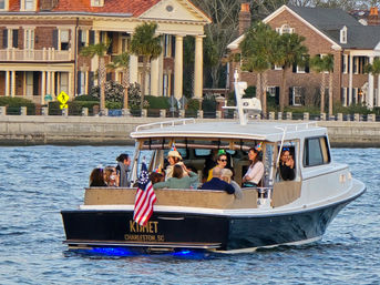 Small motor yacht with a group wearing party hats and an American flag cruising Charleston, SC harbor past historic waterfront mansions and palm trees.
