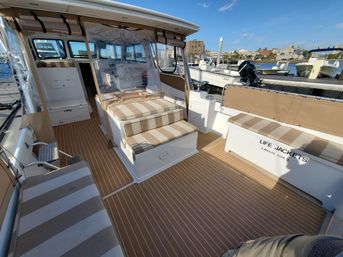 Sunlit boat deck moored at a marina with tan-and-white striped cushions on built-in benches, teak-style flooring, an enclosed helm with clear vinyl curtains, labeled life jacket storage, and other boats and harbor buildings visible at the dock.