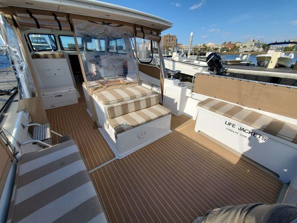 Sunlit boat deck moored at a marina with tan-and-white striped cushions on built-in benches, teak-style flooring, an enclosed helm with clear vinyl curtains, labeled life jacket storage, and other boats and harbor buildings visible at the dock.