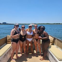 Group of five friends in swimsuits smiling and holding drinks on a sunny summer boat deck in calm blue bay waters with a shoreline and another boat in the distance