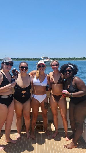 Group of five friends in swimsuits smiling and holding drinks on a sunny summer boat deck in calm blue bay waters with a shoreline and another boat in the distance