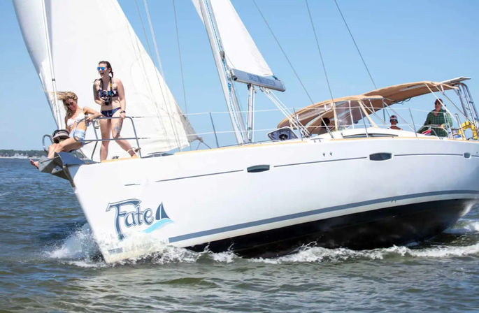 White sailboat slicing through coastal bay under a blue sky, people enjoying the sun at the bow and in the cockpit on a summer day.