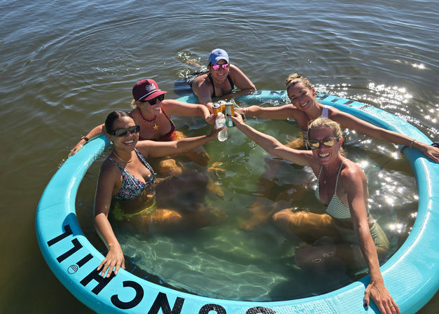 Five friends in swimsuits lounging inside a turquoise inflatable floating ring on a sunny lake, smiling and clinking drinks for a relaxed summer outing.