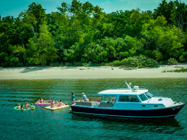 Motor yacht anchored off a sandy, tree-lined shore as a group of people relax on colorful inflatable tubes and rafts in calm blue-green water