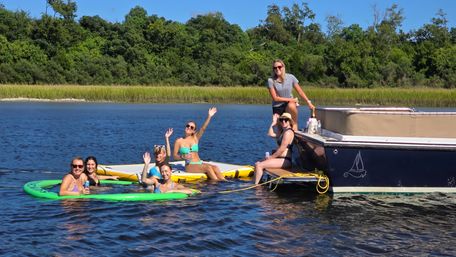 Friends waving and smiling on paddleboards and inflatable mats beside a navy pontoon boat on a sunny coastal marsh inlet