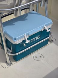 Teal and light-blue hard cooler tucked under stainless-steel seat rail on a white boat deck