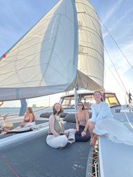 Three friends lounging on the netted deck of a catamaran under full sails during a golden-hour coastal sunset sail.