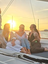 Three friends lounging on a sailboat deck at golden sunset over the water, wearing sunglasses and summer outfits, one holding a drink — relaxed sunset sail vibe.