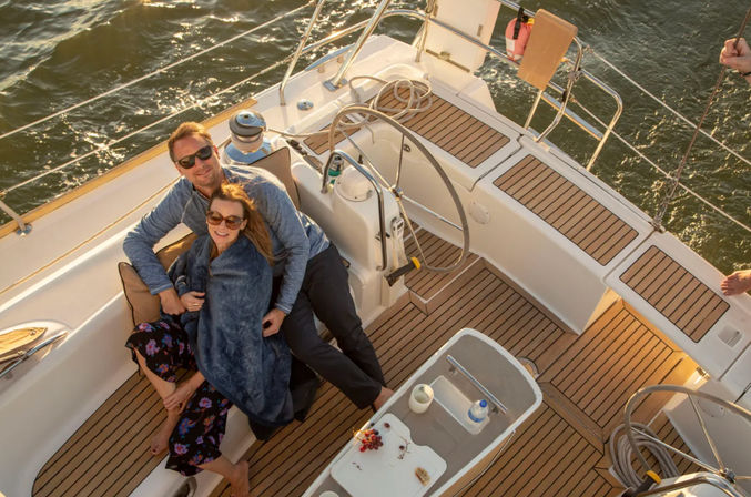Cozy couple relaxing on a sailboat's teak cockpit at golden hour, wrapped in a blanket near the helm with a small table holding snacks and a water bottle and calm open water beyond.