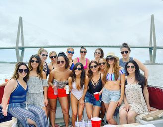 Group of friends on a boat party near a cable-stayed bridge, women in swimsuits and summer outfits smiling and holding drinks on a cloudy waterfront day