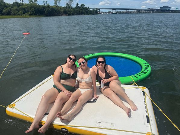 Three friends in swimsuits lounging on a white inflatable dock beside a green circular floating trampoline on a sunny river, with a distant highway bridge and tree-lined shoreline.