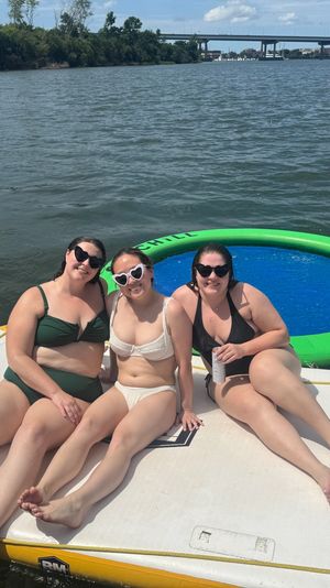Three friends in swimsuits lounging on a white inflatable dock beside a green circular floating trampoline on a sunny river, with a distant highway bridge and tree-lined shoreline.