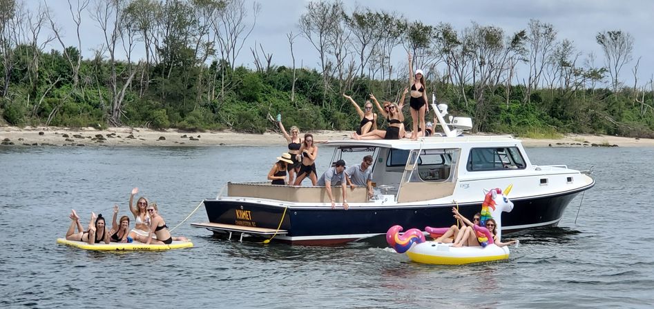 Boat party in a coastal inlet: a cabin cruiser anchored near a sandy, tree-lined shore with a group in swimsuits on deck and on inflatables — a yellow floating mat and a unicorn float.