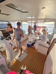 Friends relaxing inside a catamaran cabin at sunset, captain at the helm and coastal harbor waters visible through windows.