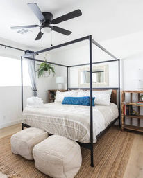 Bright airy modern bedroom with black metal canopy bed, white textured bedding and blue accent pillow, jute rug, two cream poufs, hanging fern, wooden bedside shelves and a black ceiling fan
