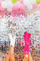 Festive photo-booth scene with two legs in white and hot-pink cowboy boots kicked up against a reflective sequin wall, pink and white balloons overhead and a neon “Let's Party” sign