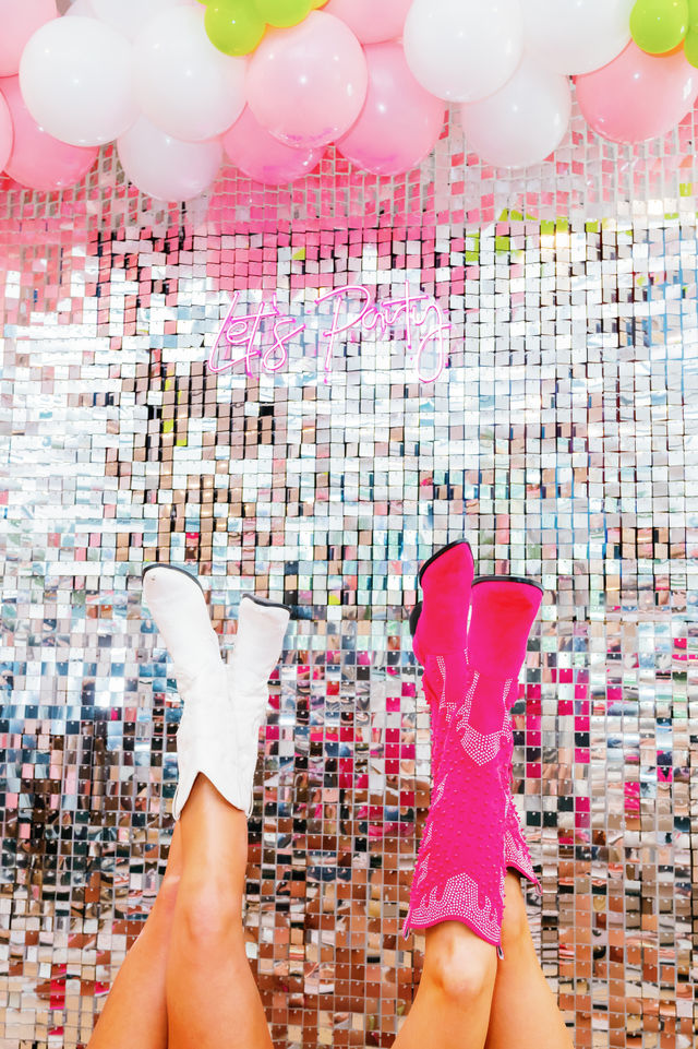 Festive photo-booth scene with two legs in white and hot-pink cowboy boots kicked up against a reflective sequin wall, pink and white balloons overhead and a neon “Let's Party” sign