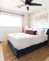 Sunlit modern bedroom with navy upholstered bed, white textured duvet, burgundy lumbar pillow, floral accent wallpaper, black ceiling fan and jute rug