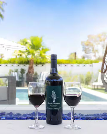 Bottle of red wine flanked by two filled wine glasses on a patio ledge with blue glass accents, overlooking a sunny backyard pool and palm trees.