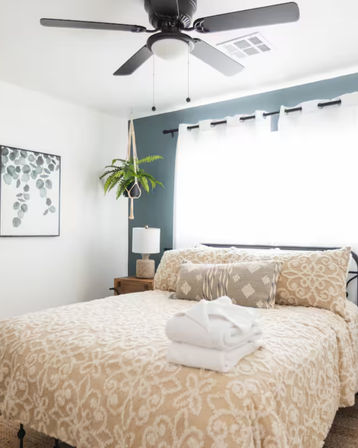 Cozy sunlit bedroom with beige patterned bedspread, stacked white towels on the bed, decorative pillows, hanging fern plant, bedside lamp, and black ceiling fan.