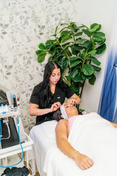 Spa esthetician in black scrubs prepping a reclining client for a facial or eyebrow treatment on a white treatment bed, surrounded by a leafy indoor plant and soft floral wallpaper in a bright treatment room.