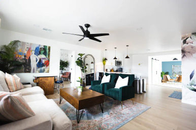 Sunlit modern open-concept living room and kitchen with emerald velvet armchairs, white sofa, wooden coffee table on a patterned rug, black ceiling fan, pendant lights, large abstract wall art and indoor plants.