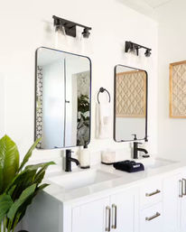 Bright modern double-sink bathroom vanity with white cabinets, quartz countertop, matte black faucets, two rounded-rectangle mirrors, glass wall sconces, woven wall art and a potted green plant