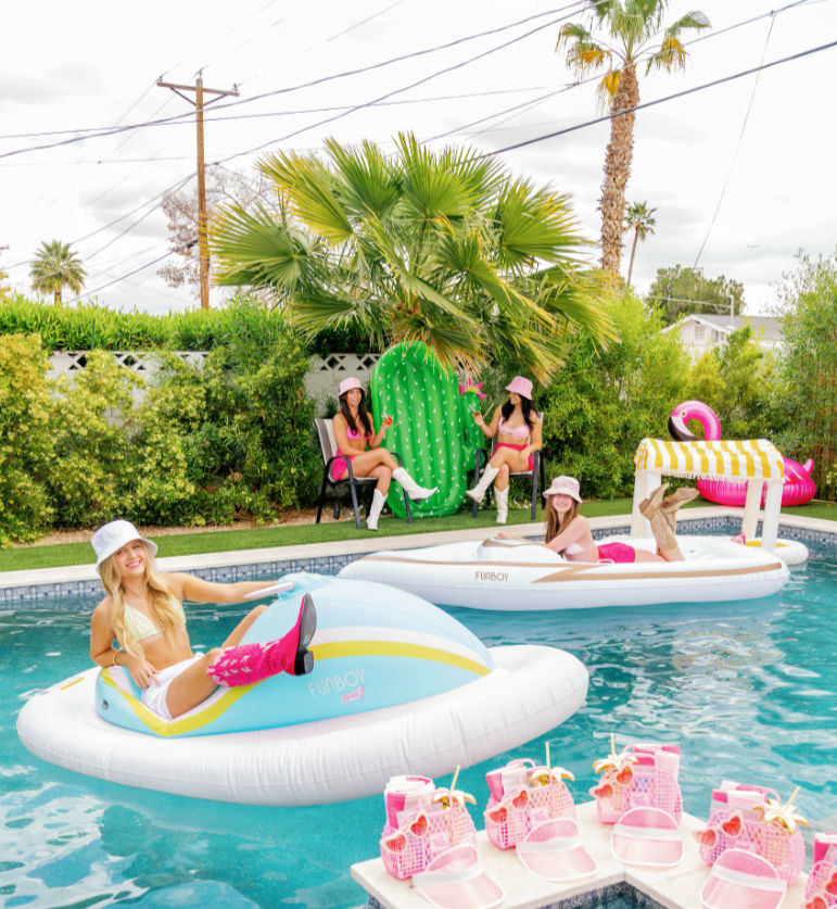 Palm-lined suburban backyard pool party with women lounging on pastel inflatable floats, a pink flamingo and cactus float, and a pool deck arranged with matching pink beach bags and sandals.