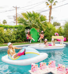 Palm-lined suburban backyard pool party with women lounging on pastel inflatable floats, a pink flamingo and cactus float, and a pool deck arranged with matching pink beach bags and sandals.