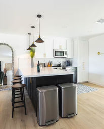 Modern open-concept kitchen with white cabinets and stainless appliances, black island with white quartz countertop, three brass-accented pendant lights over a breakfast bar with wooden stools, and two stainless steel step trash cans on light hardwood floors.