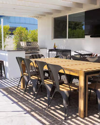 Covered backyard patio with a long wooden dining table and black metal chairs, stainless steel grill and bar cart under a sun-striped pergola.