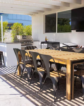 Covered backyard patio with a long wooden dining table and black metal chairs, stainless steel grill and bar cart under a sun-striped pergola.