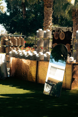 Sunny outdoor hat display under palm trees — stacks of white and tan straw hats on metallic gold counters beside a full-length mirror reflecting a balcony and greenery.