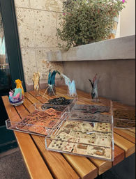 Sunlit outdoor patio table with clear acrylic trays of vintage playing cards and jars of colorful craft feathers beside a stone planter