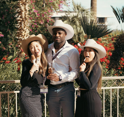 Three smiling people in cowboy hats and conference badges enjoying chocolate-covered ice cream bars on a sunny, palm-lined courtyard during an outdoor networking event