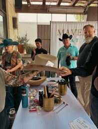 Artisan hat-shaping booth at an outdoor market with people trying on wide-brimmed cowboy hats, hat blocks and shaping tools displayed on a table.