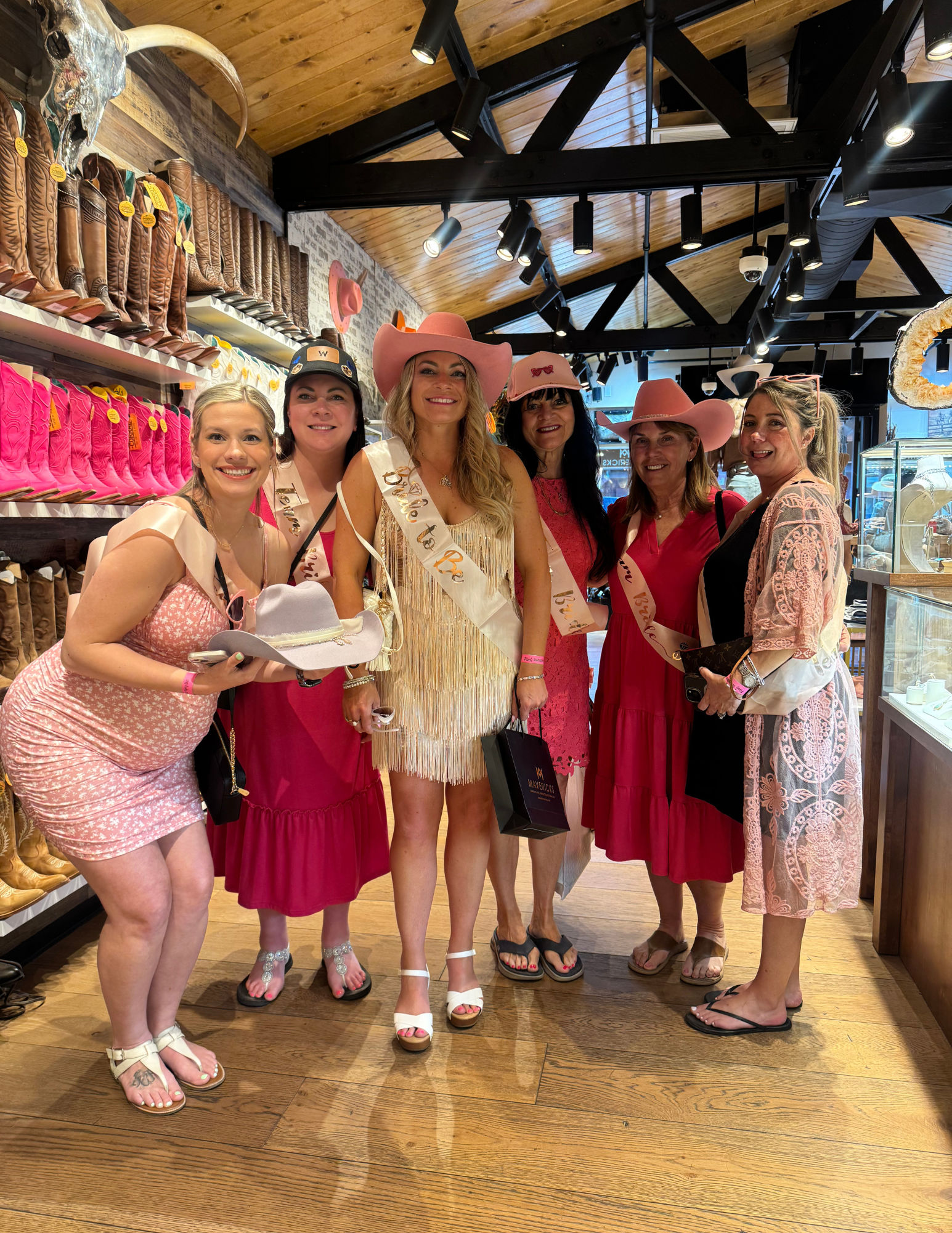 Bachelorette party of six women wearing pink cowboy hats and sashes, smiling and shopping in a western boot shop aisle lined with pink and brown cowboy boots.