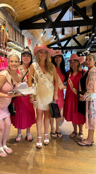 Bachelorette party of six women wearing pink cowboy hats and sashes, smiling and shopping in a western boot shop aisle lined with pink and brown cowboy boots.