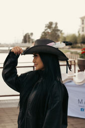 Outdoor portrait of a woman tipping a feather-adorned cowboy hat, wearing a black leather jacket and long dark hair, on a marina boardwalk with water and trees blurred in the background.
