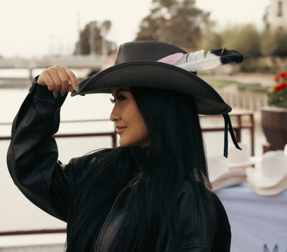 Outdoor portrait of a woman tipping a feather-adorned cowboy hat, wearing a black leather jacket and long dark hair, on a marina boardwalk with water and trees blurred in the background.