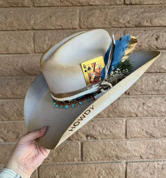 Hand holding beige felt Western cowboy hat stamped “HOWDY” on the brim, decorated with a turquoise-beaded hatband, blue and gold feathers, greenery and a tucked playing card, against a tan brick wall.