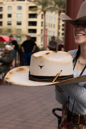 Person holding a toasted cream felt cowboy hat with longhorn emblem, leather and braided hatband, and wide brim at an outdoor market plaza