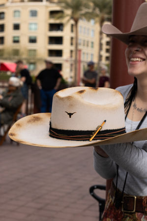 Person holding a toasted cream felt cowboy hat with longhorn emblem, leather and braided hatband, and wide brim at an outdoor market plaza