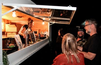 Night market food truck window with a smiling server in a wood-paneled trailer serving drinks to a small group of patrons under warm hanging lights