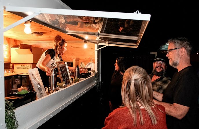 Night market food truck window with a smiling server in a wood-paneled trailer serving drinks to a small group of patrons under warm hanging lights